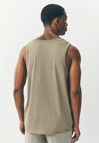 Beige ilmanhiha tank-top pehmeästä puuvillakankaasta. Ominaisuuksia ovat pyöreä kaula-aukko, siistit saumat ja rento istuvuus, nähtynä takaa.