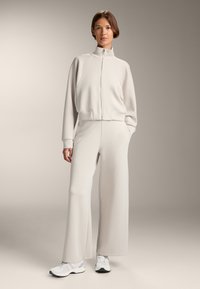 Svijetlosiva, rebrasta jakna s patentnim zatvaračem i visokim ovratnikom, uparena s odgovarajućim labavim hlačama. Model nosi bijele tenisice. Pozadina je neutralna.