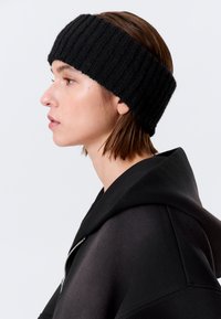 Sort ribstrikket pandebånd, brugt sammen med en sort hættetrøje med lynlås. Pandebåndet er tykt, tekstureret og sidder tæt omkring panden.