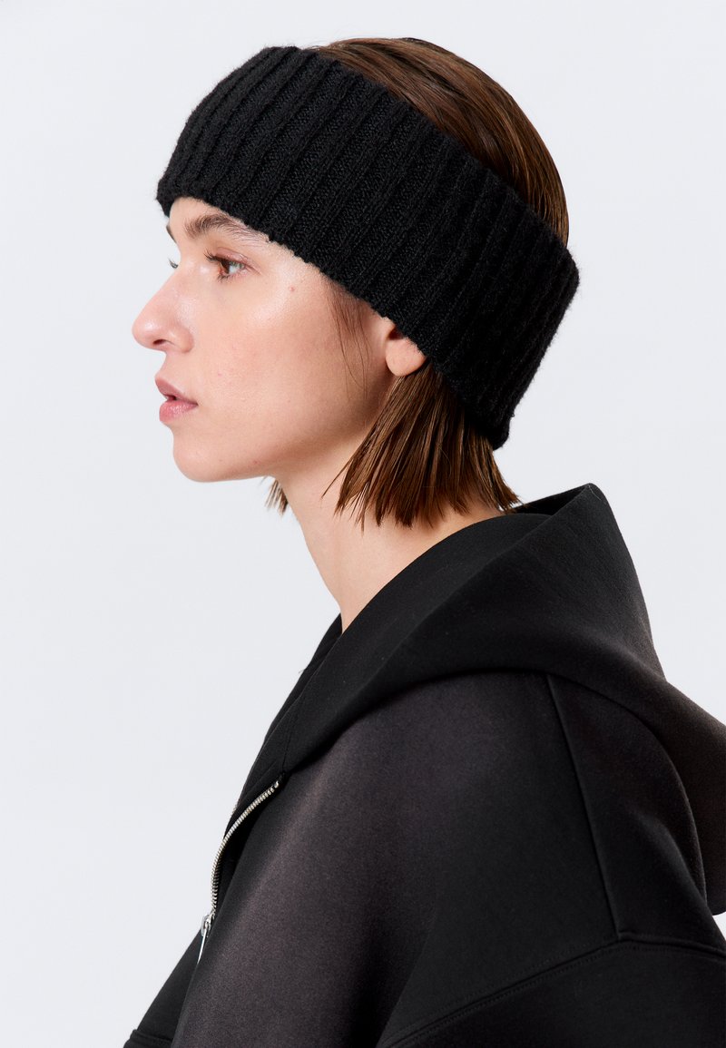 Sort ribstrikket pandebånd, brugt sammen med en sort hættetrøje med lynlås. Pandebåndet er tykt, tekstureret og sidder tæt omkring panden.