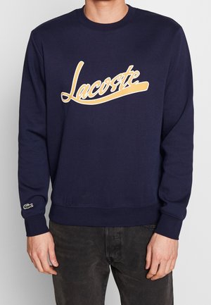 Man draagt een navyblauwe trui met gele "Lacoste" tekst over de borst en een klein groen krokodillenlogo op de manchetkant van de linker mouw.