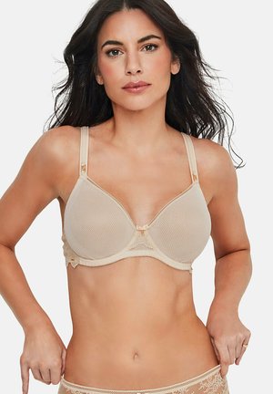 Femme aux longs cheveux foncés portant un soutien-gorge beige texturé avec des détails en dentelle, ajustant une culotte assortie en dentelle sur fond blanc.