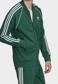 Grön polyester träningsoveralljacka med vita ränder längs ärmarna, dragkedja fram, sidofickor och rund krage.