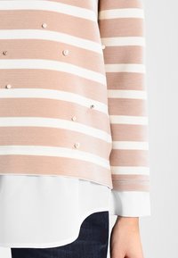 Pull rayé rose et blanc avec une finition texturée, orné de perles décoratives et d'un ourlet arrondi, porté sur une chemise blanche à plusieurs couches.