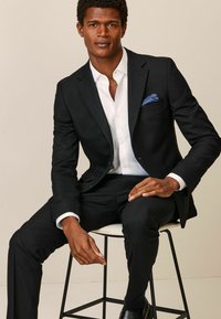 Chaqueta de traje negra sobre una camisa blanca, con un pañuelo de bolsillo azul. Corte ajustado, textura suave, con pantalones negros y zapatos elegantes.