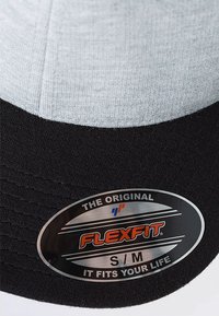 Flexfit baseball cap i ljusgrått och svart. Har en slät tygövre del, texturerad band och en metallisk oval märkesskylt.