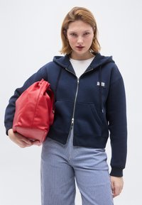 Νέα γυναίκα που φοράει μπλε ζακέτα με φερμουάρ, ριγέ παντελόνια, κρατώντας μια μεγάλη κόκκινη δερμάτινη τσάντα σε απλό ανοιχτόχρωμο φόντο.