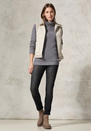 Gilet beige clair avec des accents en shearling, pull gris texturé, jean foncé et bottines beiges à semelles épaisses ; arrière-plan neutre.