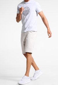 Helles graues T-Shirt mit einem schwarzen, runden Logo, kombiniert mit beigen Shorts und weißen Sneakers. Lässiger Schnitt mit kurzen Ärmeln und knielangen Shorts.