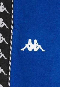 Sininen kangas sisältää valkoisen logon, jossa on kaksi istuvaa hahmoa, mustan materiaalin reunustamana, jossa on toistuva valkoisten hahmojen kuvio ja valkoinen tikkaus.