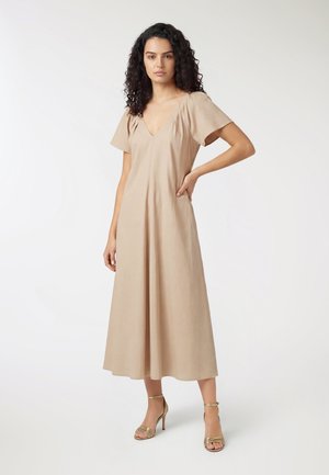 Donna con capelli ricci che indossa un vestito midi color beige con scollo a V e sandali dorati con tacco alto, in piedi con una mano sui fianchi.