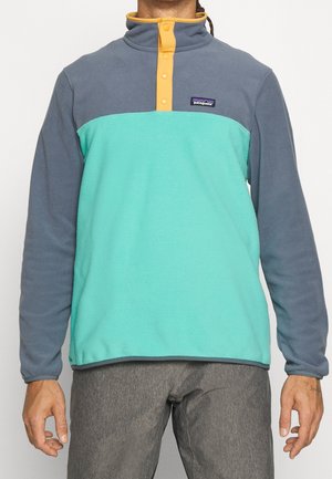 Mężczyzna ubrany w polarową bluzę dwukolorową z szarymi rękawami, turkusowym korpusem i żółtym kołnierzem na guziki, w połączeniu z szarymi spodniami.