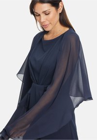Marineblaues Kleid mit transparenten, fließenden Ärmeln, das eine taillierte Brust und einen Stoffgürtel aufweist, der die Taille betont. Glatte Textur im gesamten Kleid.