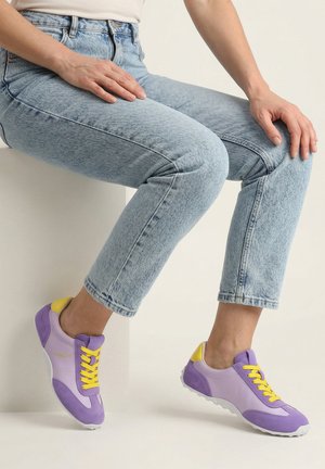 Persoon die een lichtblauwe spijkerbroek en paarse sneakers met gele veters draagt, zittend op een wit blok tegen een effen achtergrond.