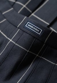 Tela azul marino con un fino patrón de cuadros blancos. Presenta una etiqueta cosida que dice "SUPERDRY" con texto blanco sobre un fondo azul oscuro.