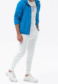 Blå lynlåstrøje med hætte, hvid T-shirt med grafisk tryk, hvide joggingbukser og hvide sneakers med synlig polstring.