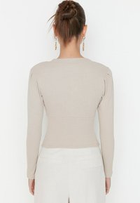 Haut beige à manches longues avec texture côtelée, silhouette ajustée et détails aux épaules. Porté avec un pantalon clair doté de poches.