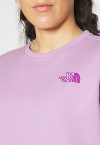 Světle fialová mikina s kulatým ribbovaným výstřihem a emblémem ve tmavší fialové, se značkou "THE NORTH FACE" na hrudi.