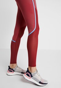 Røde leggings med blå detaljer, der har en tætsiddende design og struktureret mønster. Kombineret med hvide og grå sportssko med en pink sål.