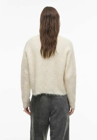 Kremasto barvni puhast pulover s kratkim krojem, z ohlapnimi dolgimi rokavi in teksturiranim zaključkom, kombiniran z temno sivimi hlačami.