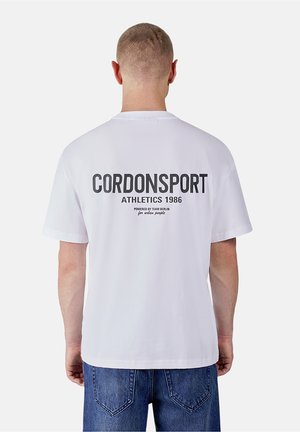 Mann mit dem Rücken zur Kamera, trägt ein weißes T-Shirt mit dem Text "CORDONSPORT ATHLETICS 1986" auf dem Rücken und blaue Jeans.