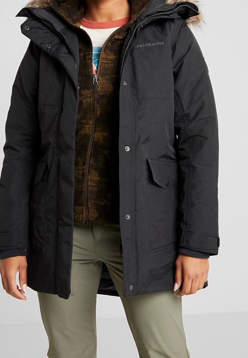 Personne portant un manteau d'hiver Didriksons noir par-dessus une veste marron à fermeture éclair et un pantalon vert, debout les mains détendues le long du corps.