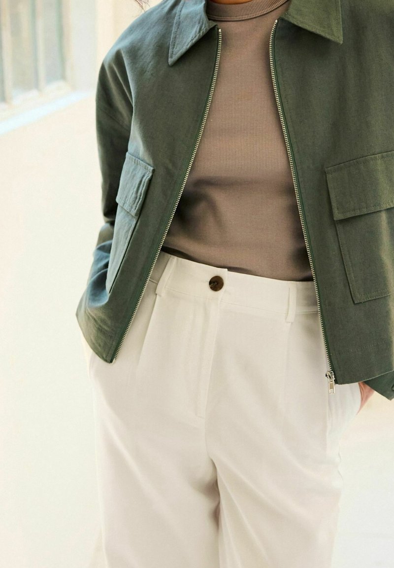 Personne portant une veste zippée vert olive sur un top côtelé marron et un pantalon blanc taille haute avec une fermeture par bouton.