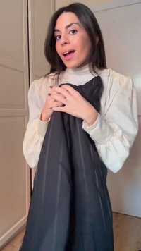 Jeune femme aux cheveux foncés et blouse blanche tenant un tissu noir à fines rayures, souriante, debout à l'intérieur près d'une porte fermée et d'une armoire.