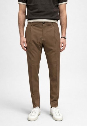 Uomo che indossa pantaloni marroni sartoriali, sneaker bianche e una camicia nera a maniche corte con bordi bianchi, in piedi davanti a uno sfondo semplice.