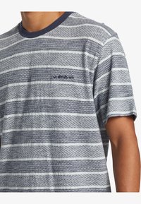 Camiseta azul a rayas con cuello redondo, que presenta una textura estampada en anchos alternos y un pequeño logo en el bolsillo del pecho.