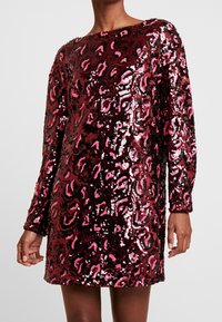 Robe à sequins avec manches longues, présentant un motif léopard en bordeaux, rose et noir. Texture lisse avec une finition brillante.