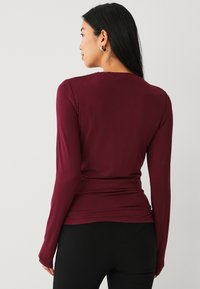 Bordeaux rood langslavige fitted top met een gladde textuur, ronde halslijn en subtiele plooien aan de achterkant, gecombineerd met zwarte onderkleding.