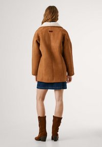 Giubbotto in suede marrone con colletto in shearling bianco, maniche lunghe e dettagli sulle cuciture posteriori, indossato sopra una gonna di jeans e stivali marroni al ginocchio.