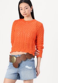 Maglione lavorato a maglia arancione con un motivo diamantato testurizzato, lunghezza crop e maniche lunghe abbinato a pantaloni shorts in denim strappati e una cintura marrone con borchie.