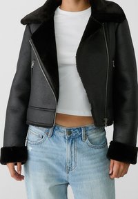 Giacca di pelle nera tagliata con colletto e polsini in pelliccia sintetica, chiusura con zip e finitura texture, abbinata a jeans in denim azzurro chiaro.