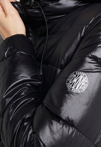 Close-up van een persoon die een glanzende zwarte pufferjack draagt met een circulaire logo-patch van Donna Karan New York op de mouw.