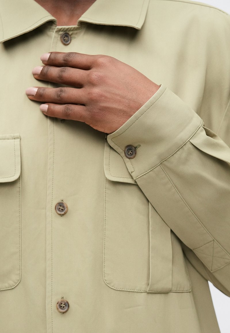 Olivgrön lättviktig skjorta med krage, framfickor och bruna knappar. Slät textur och avslappnad passform, med detaljer vid ärmsluten.