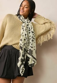 Écharpe légère et à motifs en crème avec des taches noires, drapée autour du cou. Portée avec un pull en tricot à franges et une jupe noire évasée.