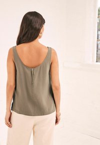Donna con lunghi capelli scuri indossa un top senza maniche color oliva con zip sul retro e pantaloni crema, in piedi all'interno vicino a una finestra.