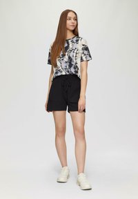 Shorts noirs avec une taille à cordon, associés à un t-shirt tie-dye dans des nuances de gris et de crème. Le modèle porte des baskets de couleur claire.