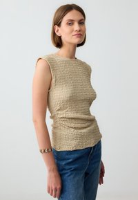 Beige textuur mouwloos topje met een ronde halslijn; gecombineerd met blauwe denim jeans. Geaccentueerd met een multi-stenen armband om de pols.