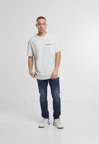 Camiseta de algodón gris con cuello redondo y mangas cortas, que presenta un pequeño gráfico en el lado izquierdo; combinada con jeans azul oscuro y zapatillas.