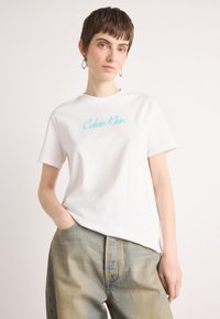 Hvit bomull t-skjorte med turkise "Calvin Klein"-logo, klassisk rund hals, korte ermer og avslappet passform, kombinert med lyse denimbukser.