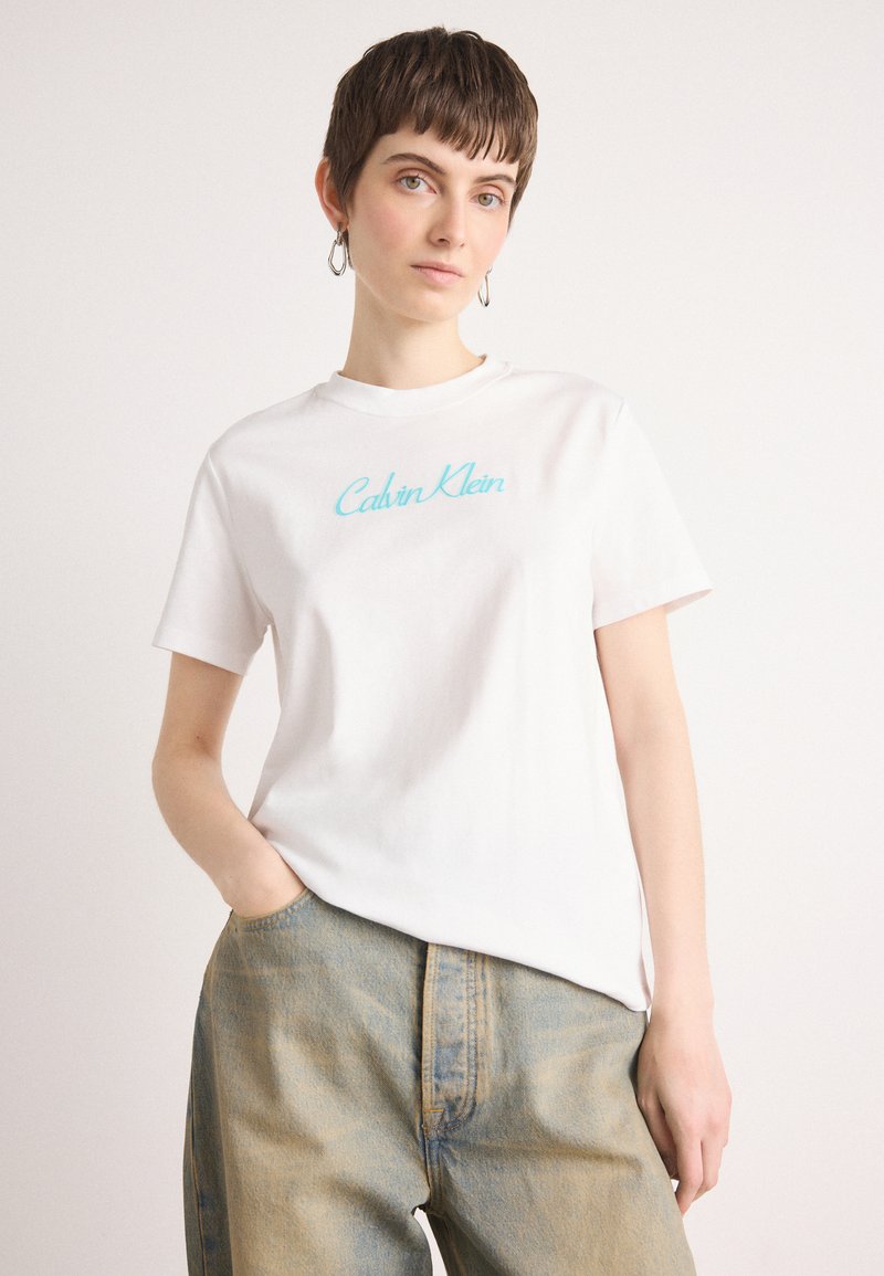 Hvit bomull t-skjorte med turkise "Calvin Klein"-logo, klassisk rund hals, korte ermer og avslappet passform, kombinert med lyse denimbukser.