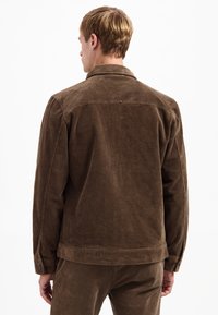 Bruine corduroyjas met een gestructureerd oppervlak, voorzien van een puntige kraag en knoopmanchetten. De achterkant toont een horizontaal ribbelpatroon.