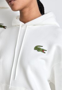 Bílá mikina s kapucí, která má na levé straně hrudi zelené vyšívané logo krokodýla, bílé stahovací šňůrky a jemnou texturu.