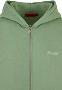 Veste à capuche zippée verte en tissu doux. Elle dispose d'une fermeture éclair en métal, d'une grande capuche et d'un petit logo "Interdit" en couleur claire sur le devant.