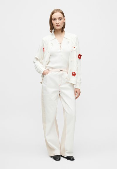 Hvit strikket cardigan med røde og rosa blomsterdekorasjoner, kombinert med kremfargede vide bukser med lyserosa søm. Svart fottøy.