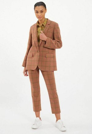 Costume à carreaux marron et rouge avec une veste structurée et un pantalon ajusté, associé à une chemise à motif floral et des sneakers blanches.