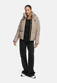 Beige puffjacka med huva, med en framdragkedja och två fickor. Modellen bär den över en svart topp och svarta vida byxor.
